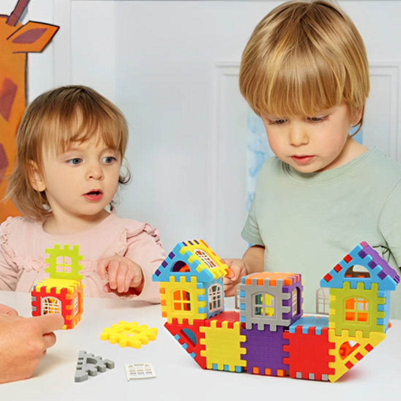 Deux jeunes enfants de 2-4 ans accompagnés par un adulte découvrant BuildCréa autour d'une table. Petites maisons colorées jaune, rouge, bleu et violet avec fenêtres blanches. Apprentissage collaboratif et guidance bienveillante pour développer la confiance en construction.