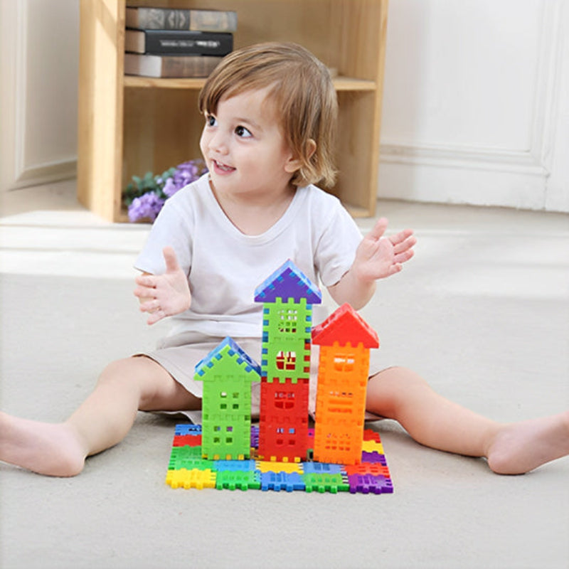 Enfant de 2-3 ans assis au sol créant avec fierté ses premières tours BuildCréa. Structure colorée de trois maisons aux toits triangulaires rouge, jaune, vert et bleu sur tapis moelleux. Développement de la motricité fine et de la créativité dans un environnement familial chaleureux.